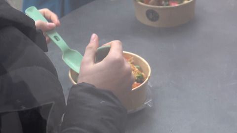 Nahaufnahme eines Gerichts in einer Pappschale. Die Schale ist mit verschiedenen Zutaten gefüllt und es steigt Dampf auf. Eine Hand ist sichtbar, die einen kleinen Behälter mit Soße hält.