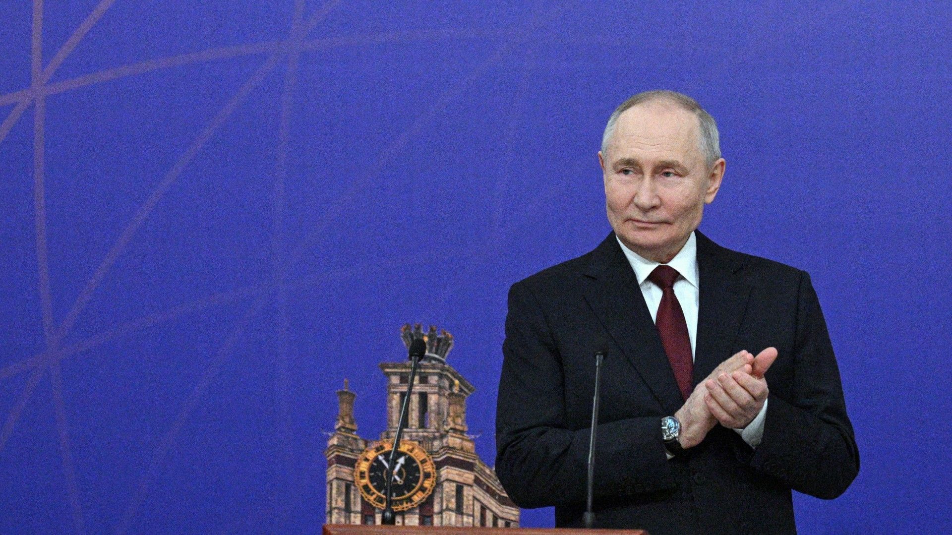 Putin n dunklem Anzug und roter Krawatte steht vor einem blauen Hintergrund mit einer stilisierten Uhr. Er applaudiert und trägt eine Armbanduhr am linken Handgelenk.