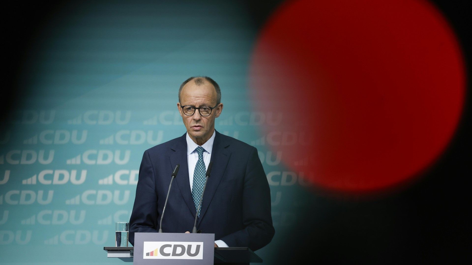 Friedrich Merz steht an einem Rednerpult bei einer Pressekonferenz der CDU. Im Hintergrund ist das CDU-Logo zu sehen, während Merz in einem formellen Anzug spricht. Ein unscharfes rotes Licht ist im Vordergrund sichtbar.